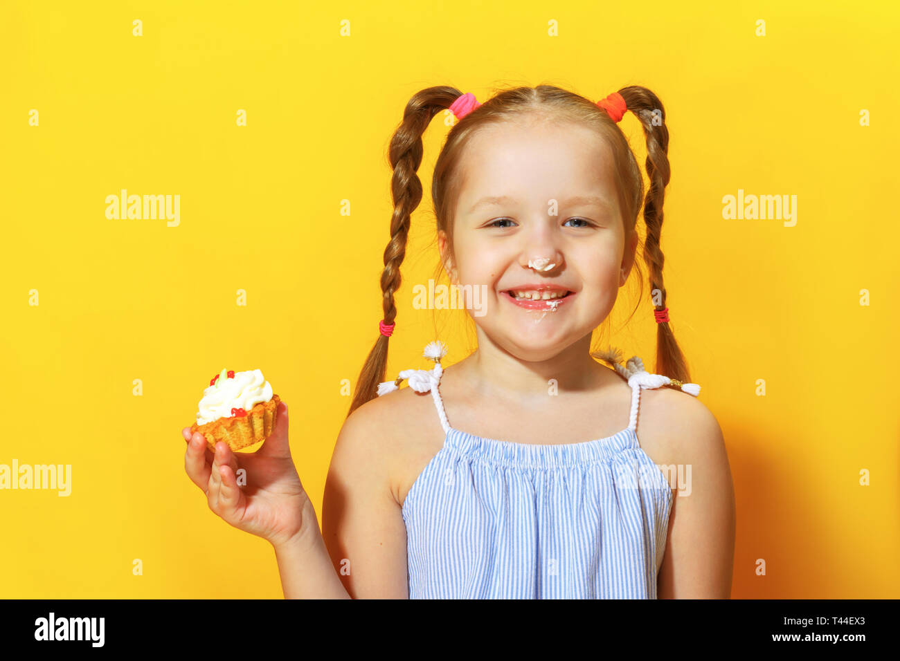 Closeup portrait of a cheerful little sweet tooth girl on a yellow background. The child smeared his nose with cream and holding a cake in his hands. Stock Photo