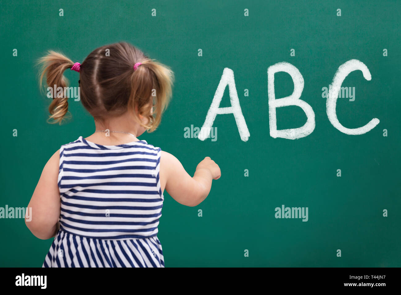 Rear View Of Schoolgirl Writing ABC Alphabet On Green Board Stock Photo