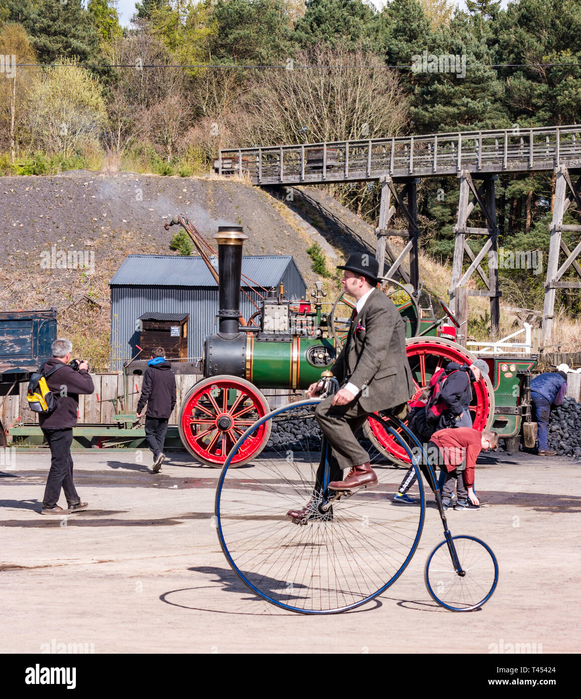Beamish Museum, Beamish, Durham County, England, United Kingdom, 13 April 2019. Beamish Steam Day: Man in vintage costume riding a penny farthing bicycle with a vintage Clayton & Shuttleworth steam traction engine on display at Beamish Living Museum Stock Photo
