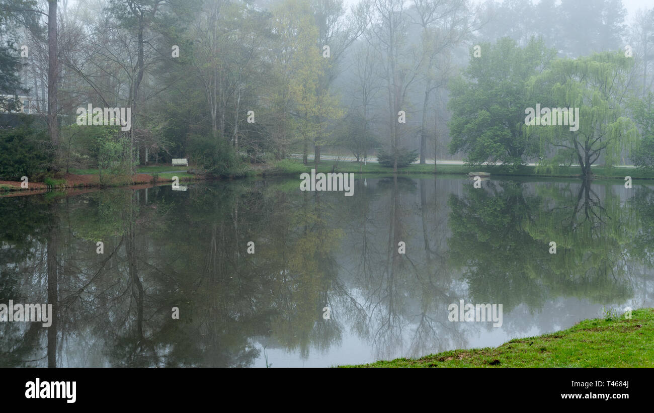 Reflection of bench and trees on pond on foggy morning Stock Photo