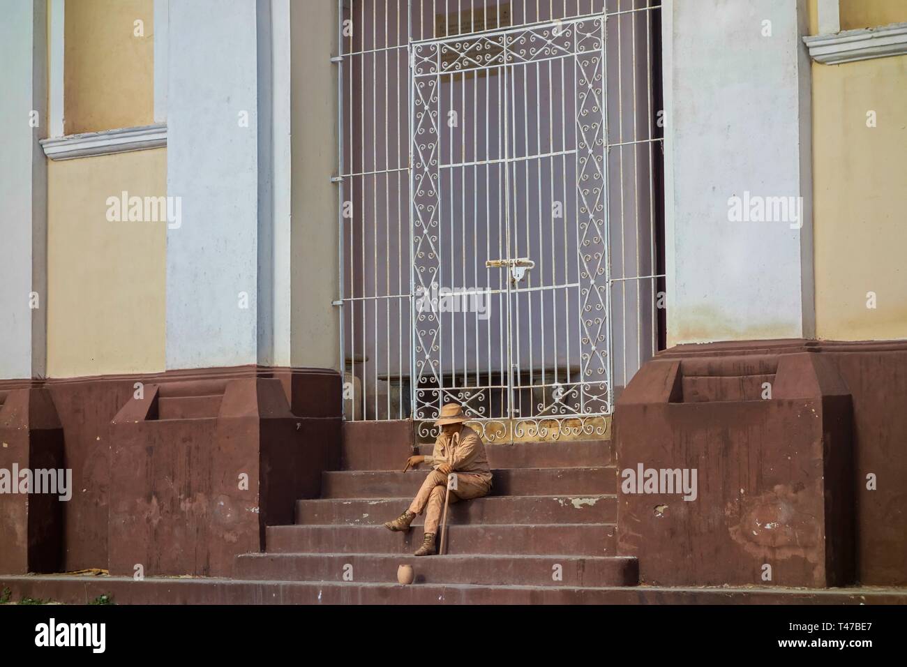 Young Man Busker with Begging Bowl covered in Paint sitting on stairs of Iglesia y Convento de San Francisco Church in Trinadad, Cuba Stock Photo