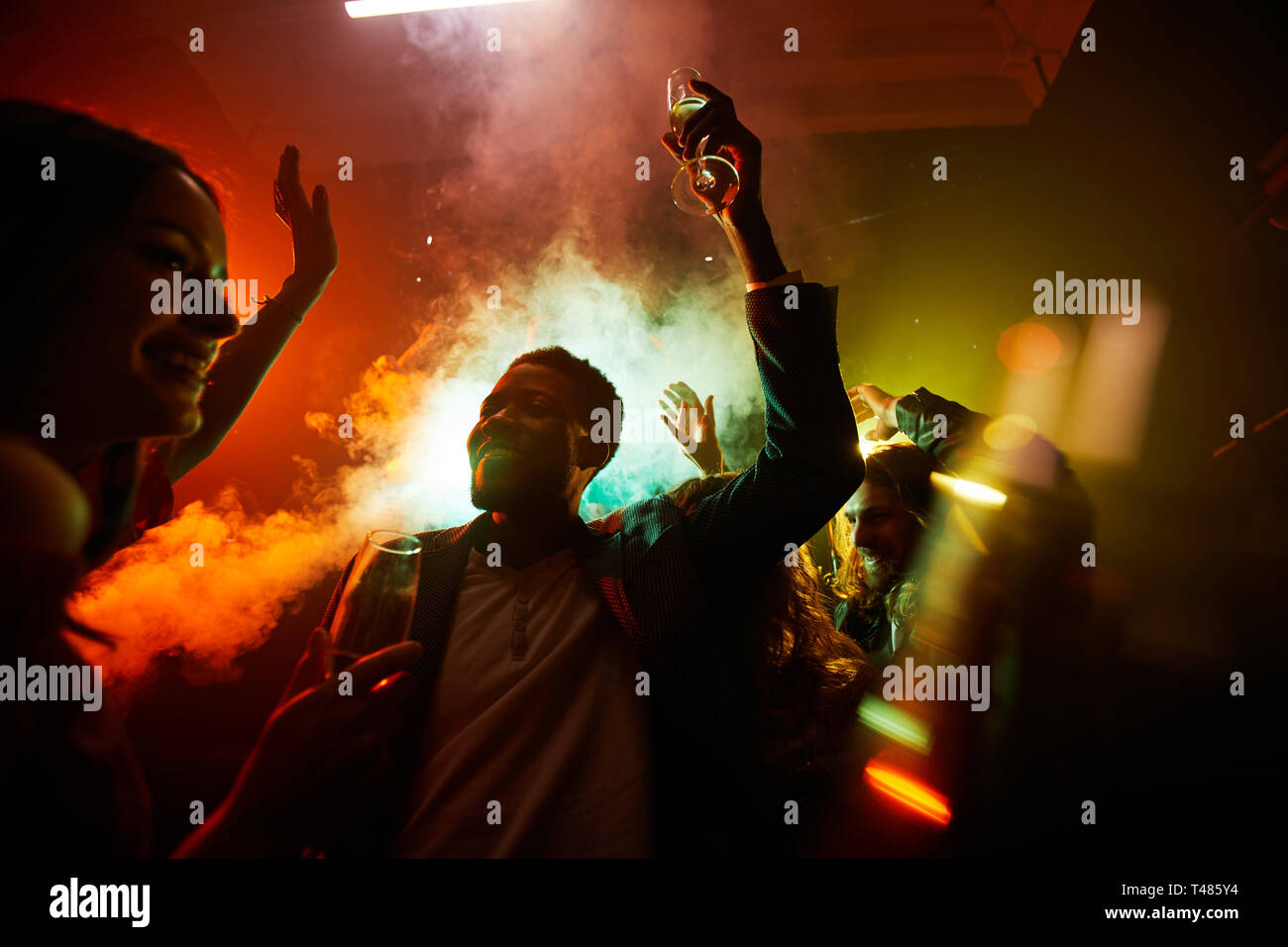Crowd of cheerful hilarious young multiethnic people drinking alcohol and dancing in smoke at noisy party Stock Photo