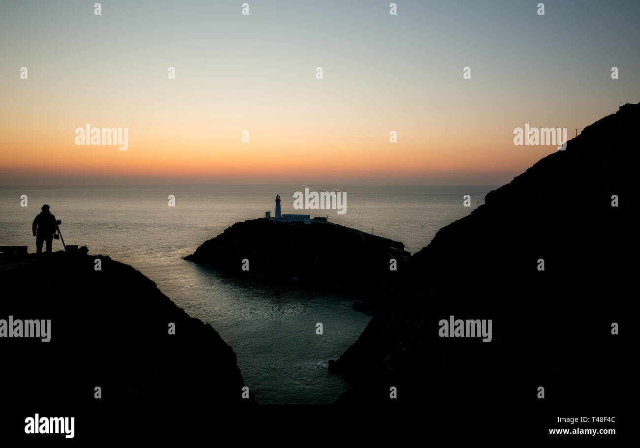 View of South Stack Lighthouse almost as a silhouette at dusk with solitary figure of photographer silhouetted on hillside taking evening photographs. Stock Photo