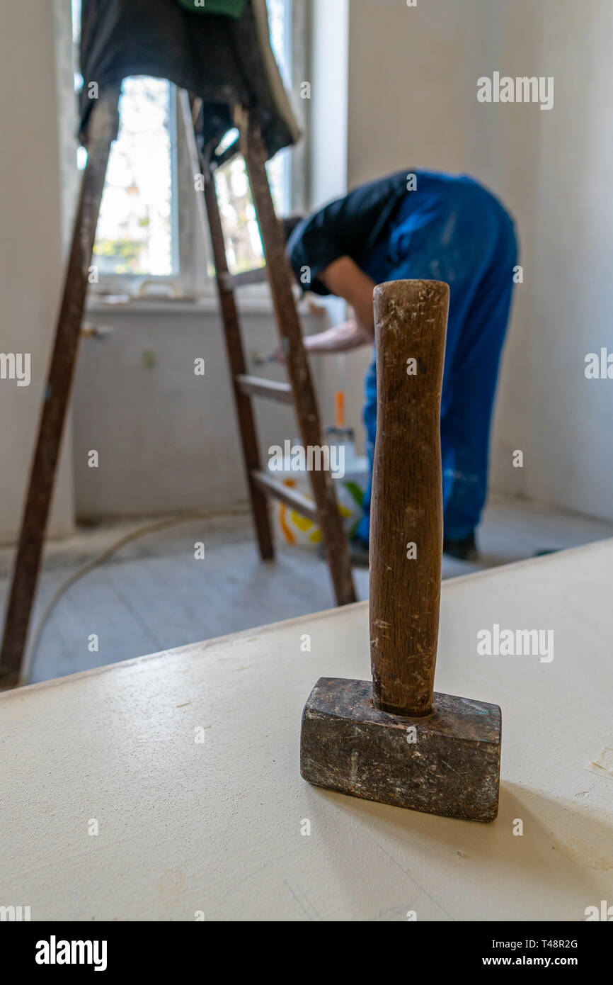 Hammer with Wooden Grip Upside Down on a Table with Worker Bending next to Ladder in Blue Colored Dungarees Trousers at Background in a Working Room Stock Photo