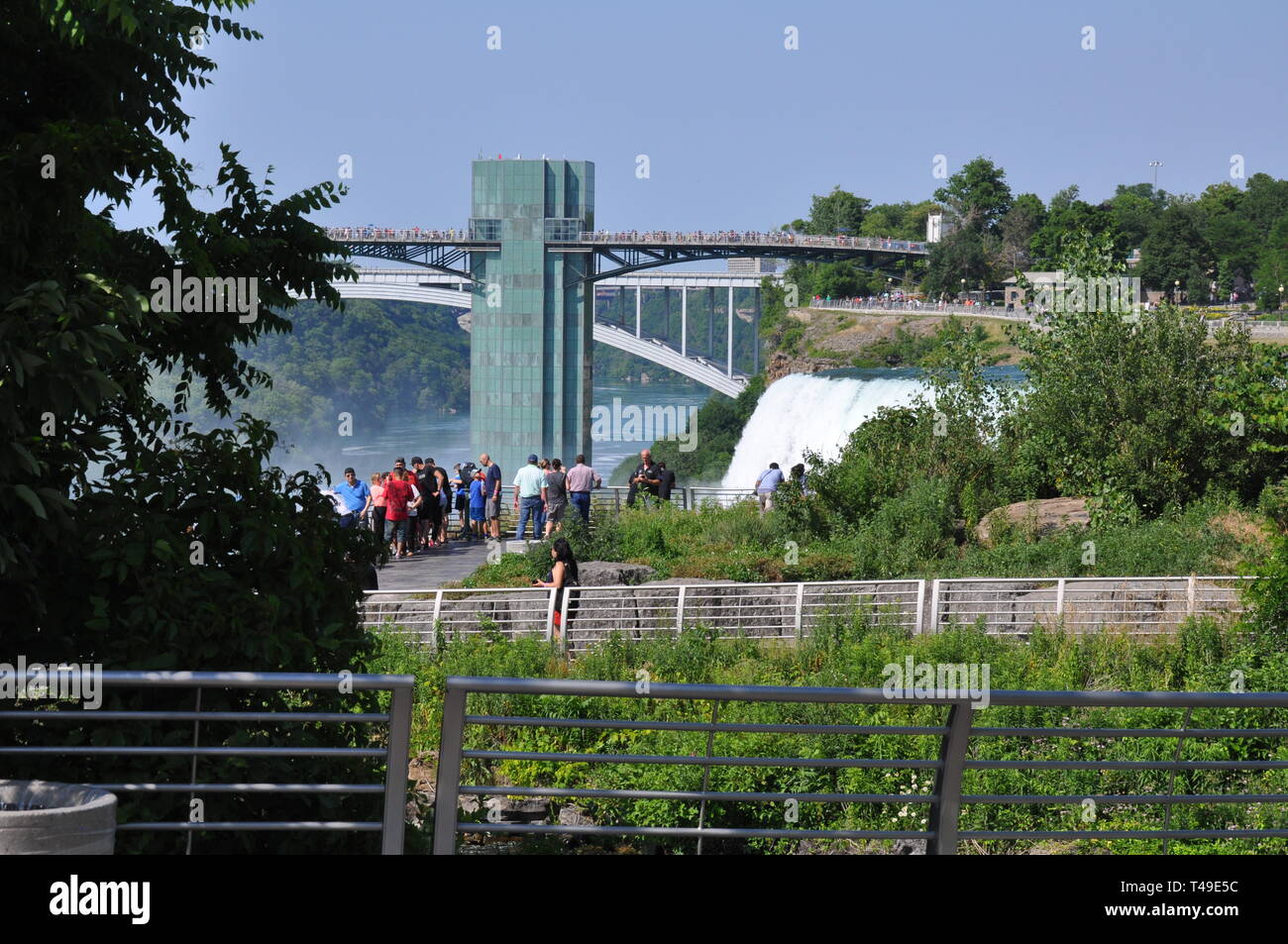 A view of Tourists at the top of the American Falls with the Rainbow Bridge and Observation tower in the background at Niagara Falls, United States Stock Photo