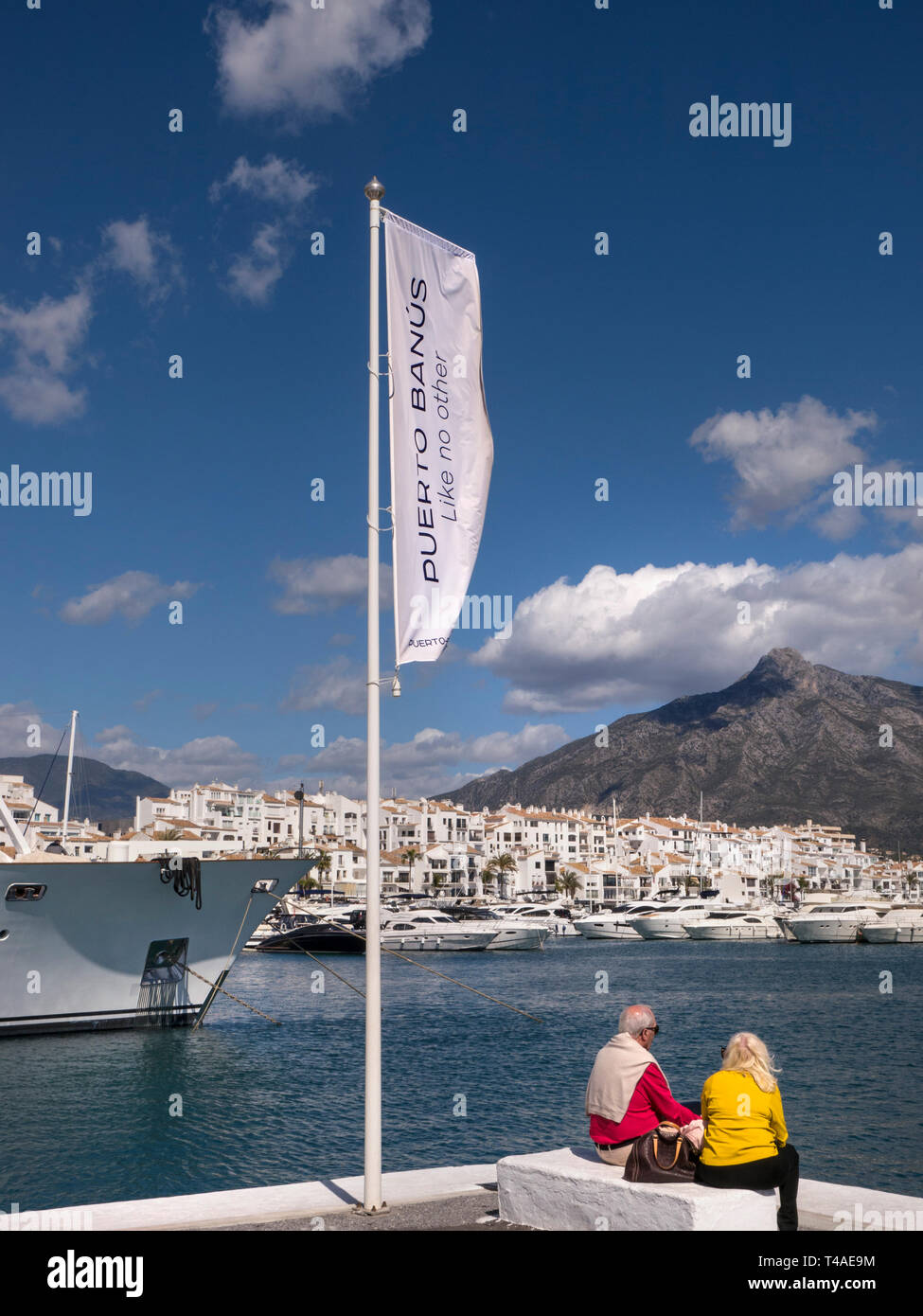 Puerto Banus Banner flying in breeze with mature holiday couple enjoying overview of super yachts of Puerto Banus marina Marbella Costa del Sol Spain Stock Photo