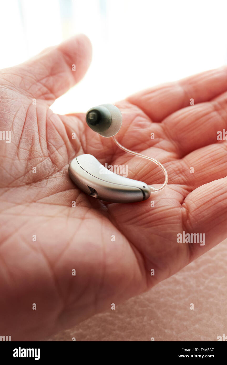 A deaf elderly person's hand holding their new Receiver-in-Canal (RIC) digital hearing aid close up. Stock Photo
