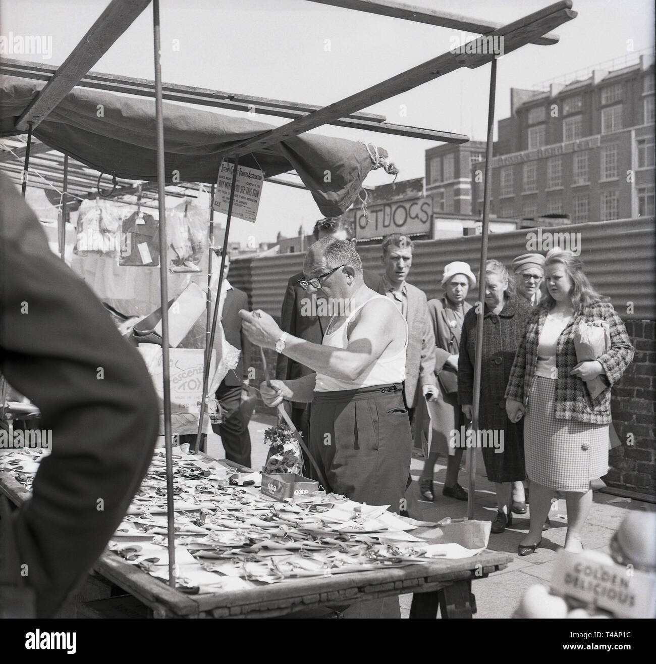 1960s, historical, male stall-holder wearing a sleeveless vest sorting out his stock of braces or trouser suspender belts on his stall,  with local people looking on, in Stepney, East London, London, England, UK Stock Photo