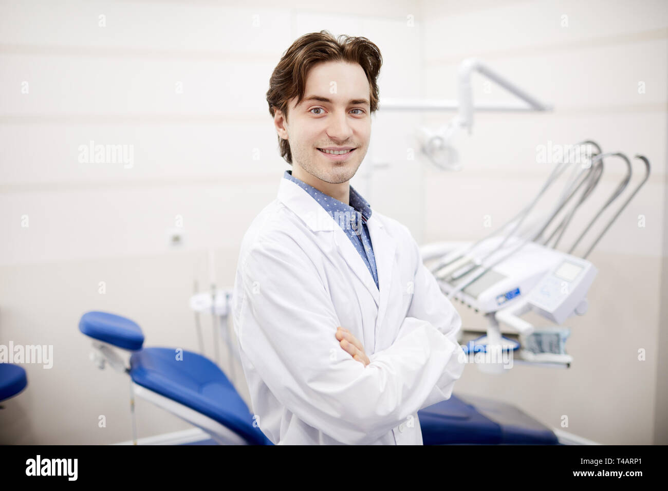 Waist up portrait of smiling young dentist posing in office next to dental chair, copy space Stock Photo