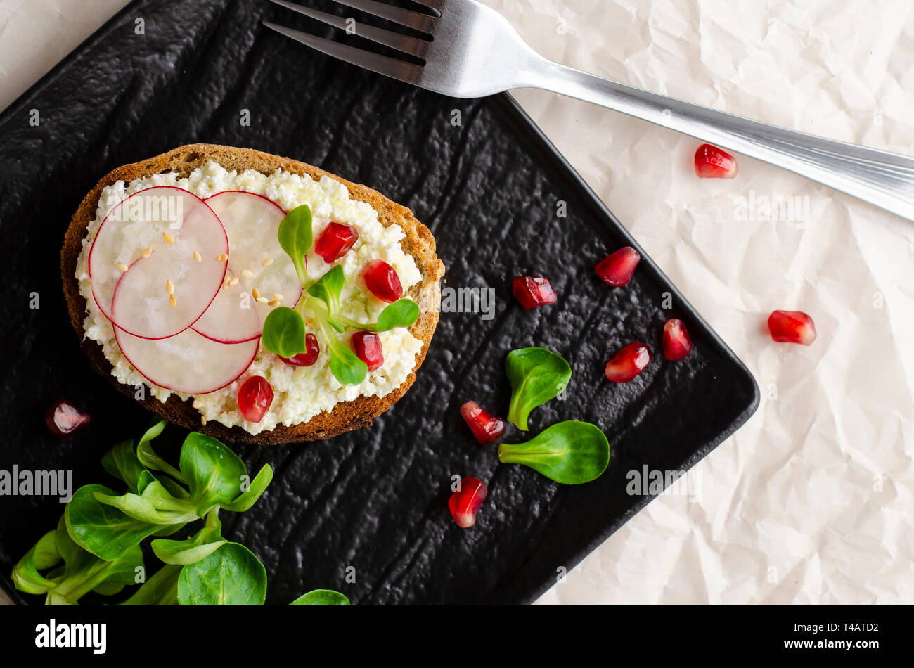 Toast with cottage cheese and smashed avocado, radish, corn salad plant and pomegranate seeds on black plate. Top view. Copy space. Healthy eating and Stock Photo