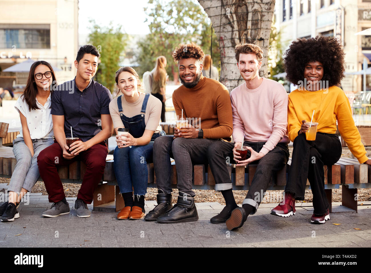 Six trendy millennial friends sitting on a wall by a fountain, drinking cold brews smiling to camera Stock Photo