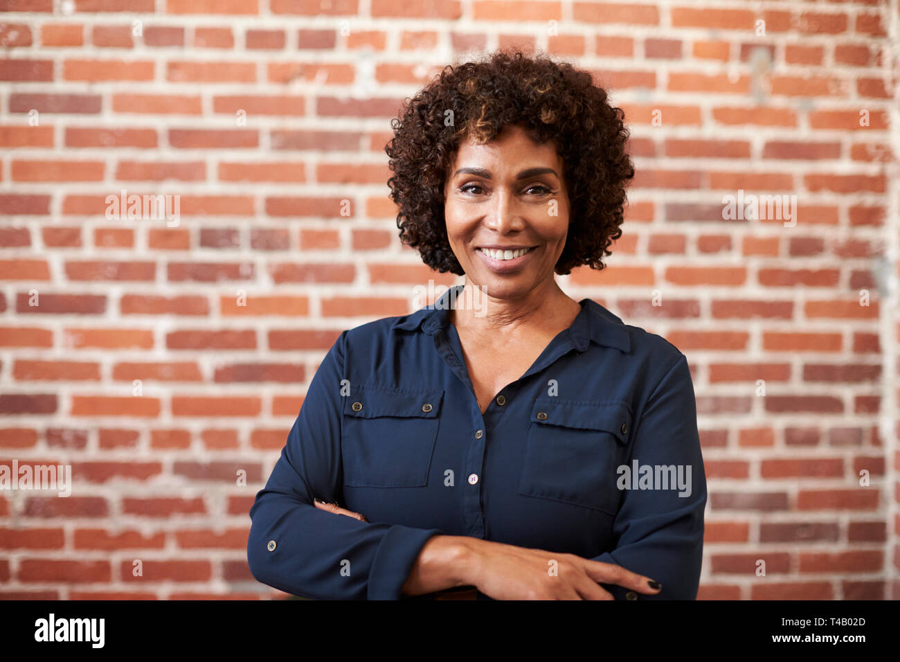 Portrait Of Smiling Mature Businesswoman Standing Against Brick Wall In Modern Office Stock Photo
