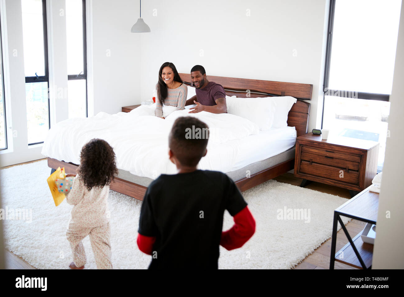 Children Running Into Parents Bedroom With Gift And Card In Bed To Celebrate Mothers Day Or Birthday Stock Photo