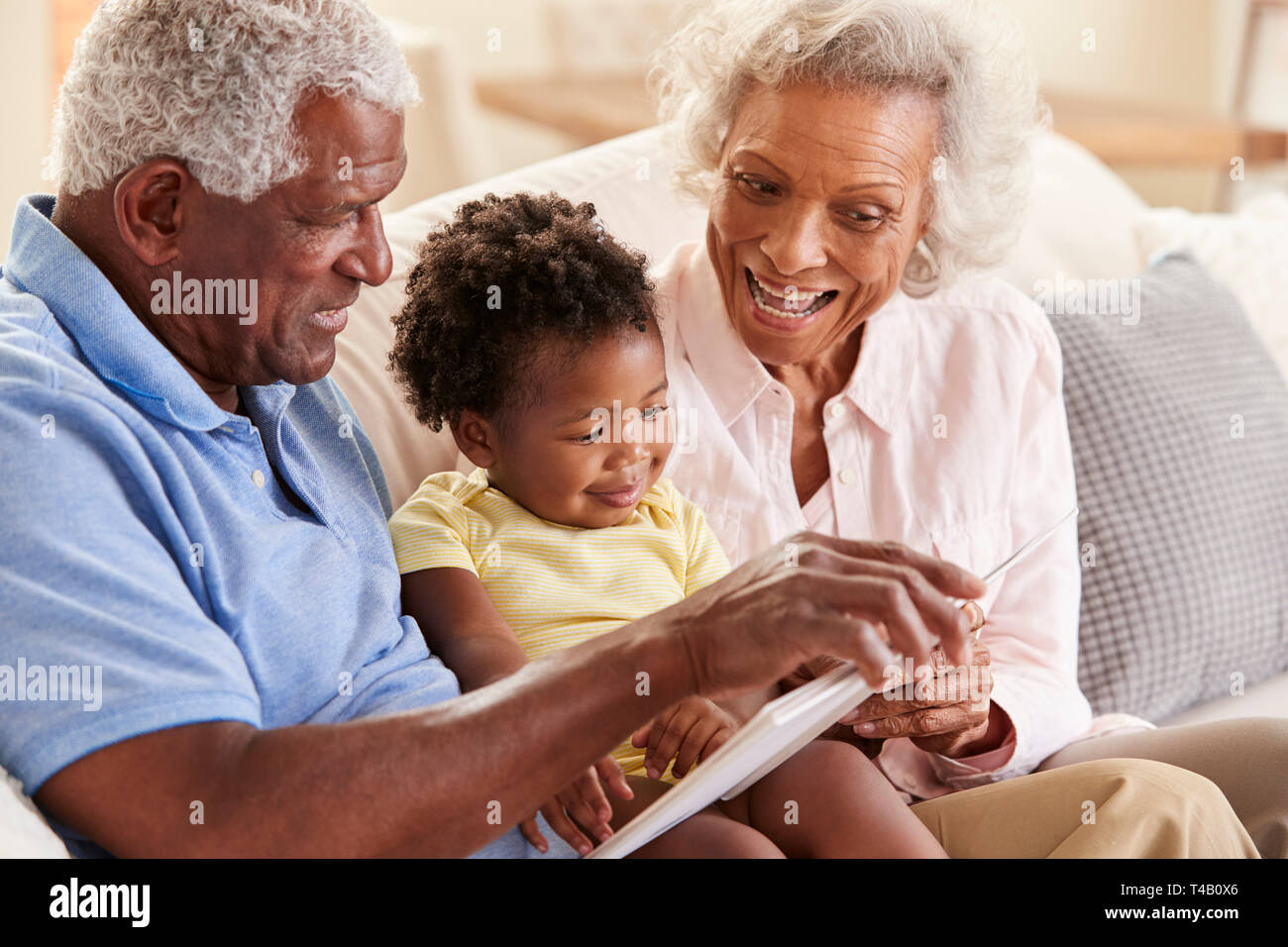 Grandparents Sitting On Sofa At Home Reading Book With Baby Granddaughter Stock Photo