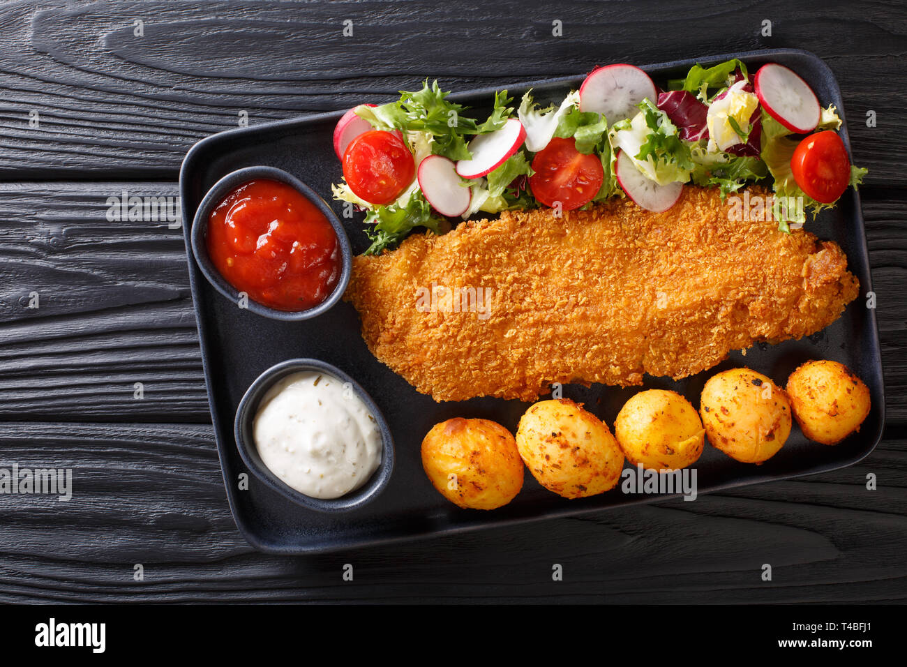 main course of cod fillet in breading with a garnish of new potatoes and fresh vegetable salad close-up on a plate on the table. Horizontal top view f Stock Photo