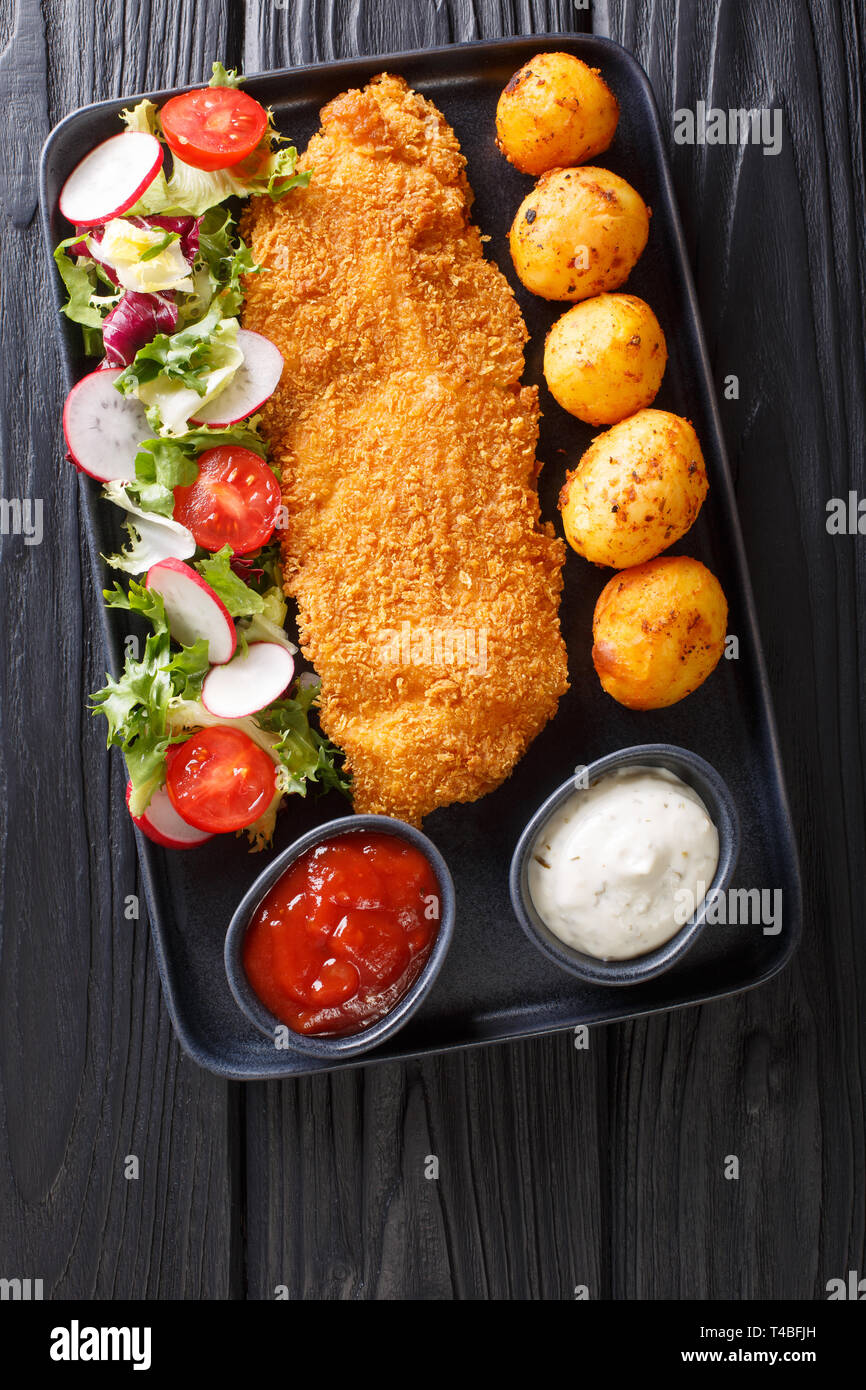main course of cod fillet in breading with a garnish of new potatoes and fresh vegetable salad close-up on a plate on the table. Vertical top view fro Stock Photo