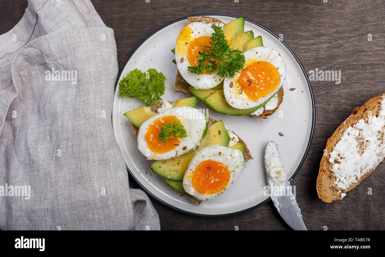 Tasty toast with cream cheese, avocado and eggs on white plate with knife and napkin at wooden background. Concept of breakfast with eggs Stock Photo