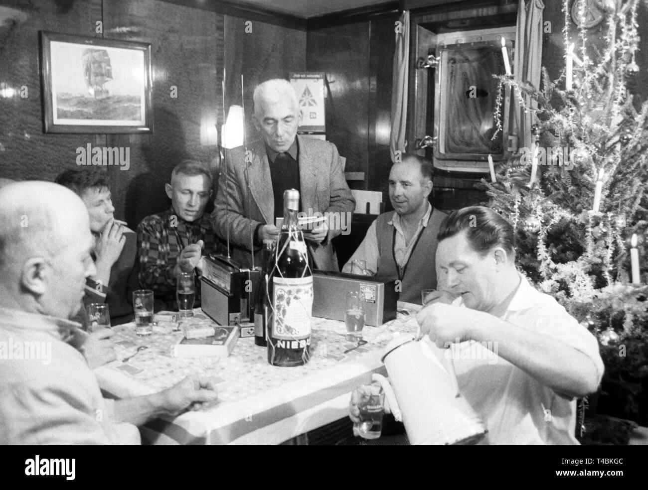 The crew of the oldest German lightship 'Bremen' sits together on 16 December 1963 during a christmas dinner in the captain's room. Captain Grelle (centre) holds a speech while Jürgens, the cook (right), pours the christmas punch. | usage worldwide Stock Photo