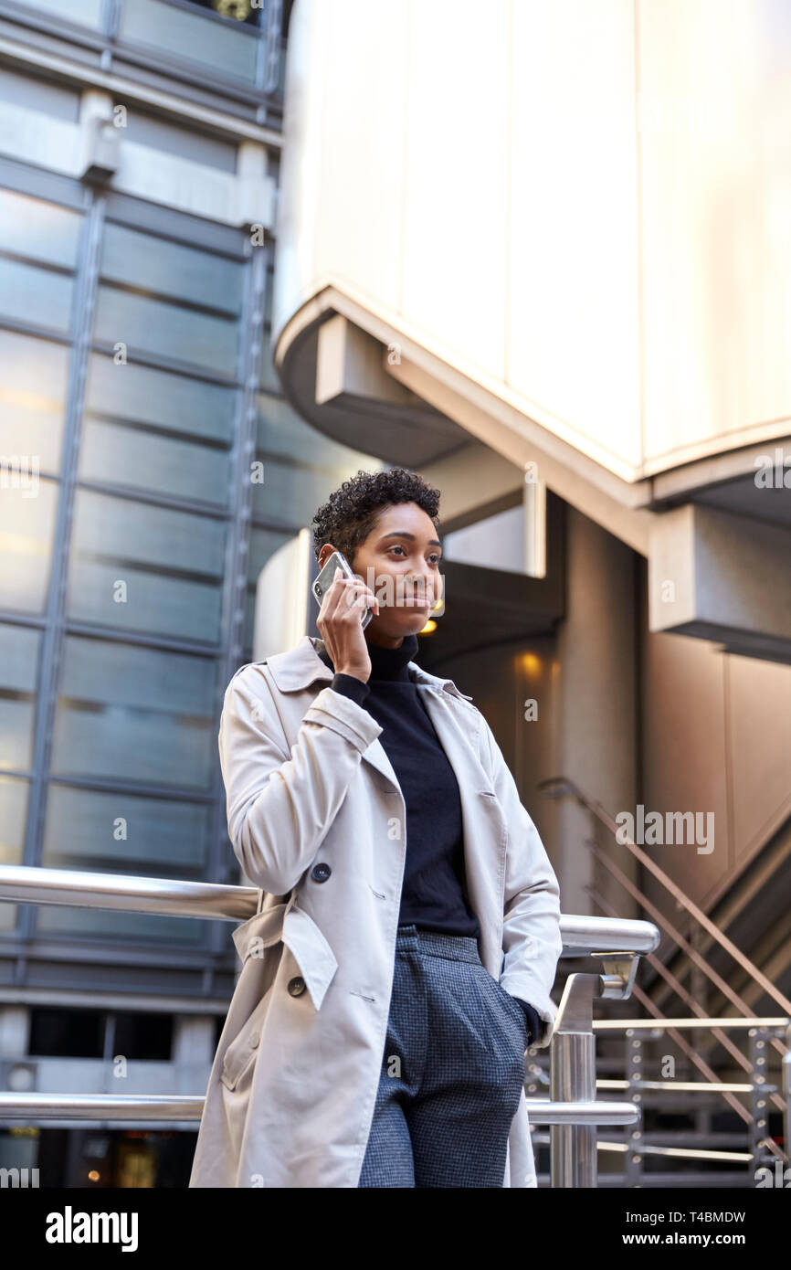 Fashionable young black businesswoman standing against handrail in the city talking on smartphone, vertical Stock Photo