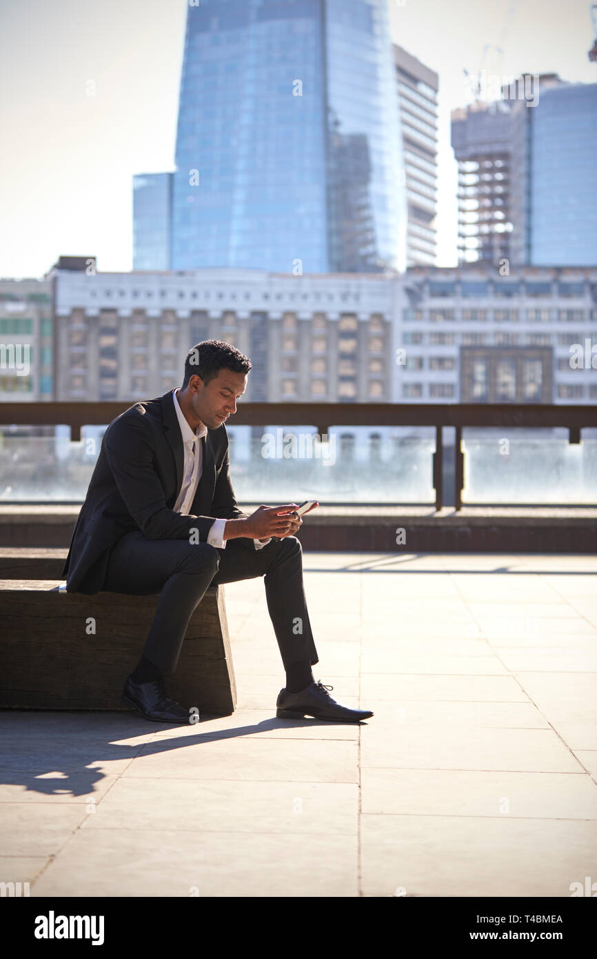 Millennial black businessman wearing black suit and white shirt sitting on the Thames embankment, London, using smartphone, vertical Stock Photo