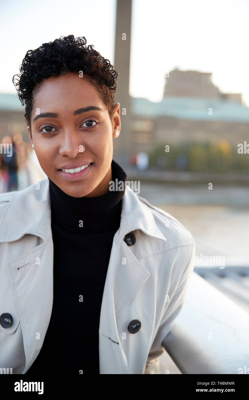 Young black woman wearing a turtleneck sweater and a mackintosh standing on Millennium Bridge, London, looking to camera smiling, vertical Stock Photo