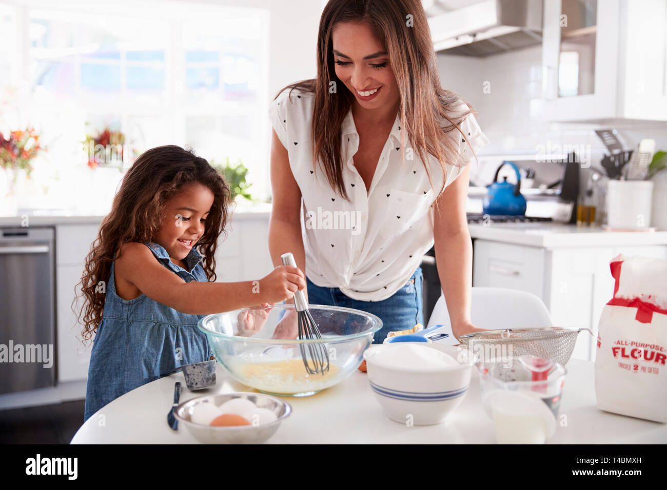 Young Hispanic girl making cake in the kitchen, overseen by her mum, waist up Stock Photo
