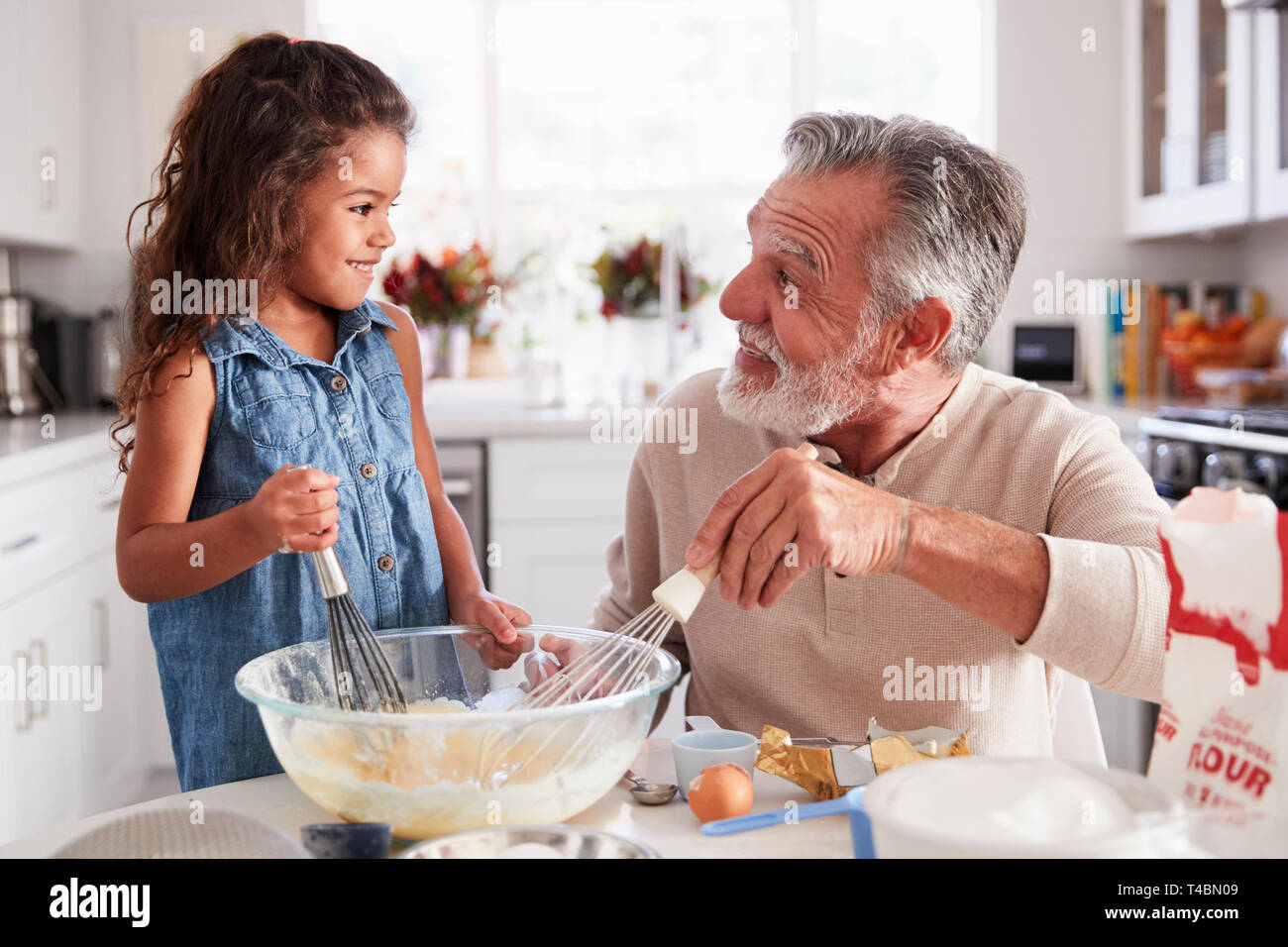 Young girl and grandfather making cake mixture at the kitchen table, smiling at each other, close up Stock Photo