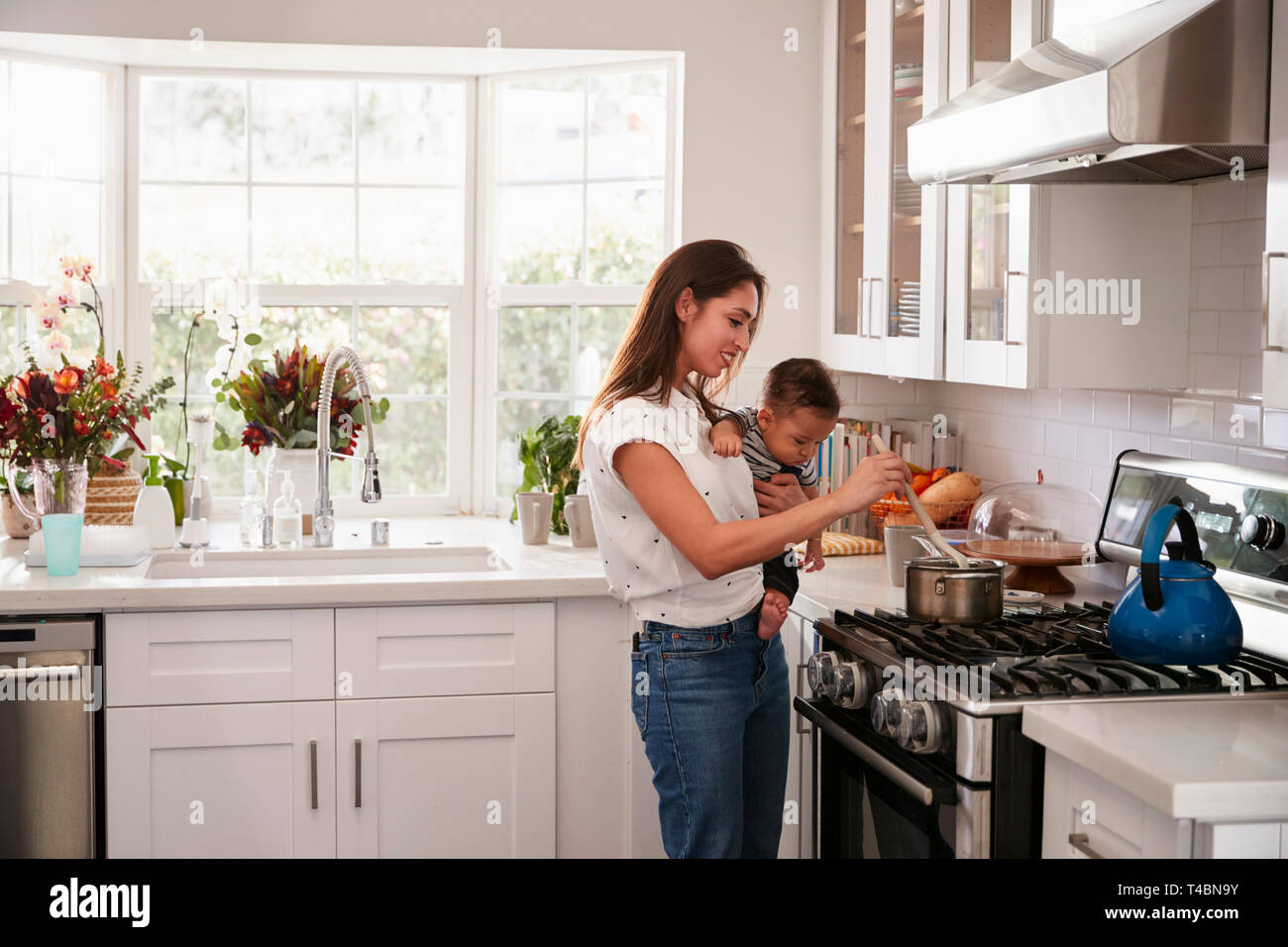 Multitasking mum holding her young baby while she makes food at the hob in her kitchen, side view Stock Photo