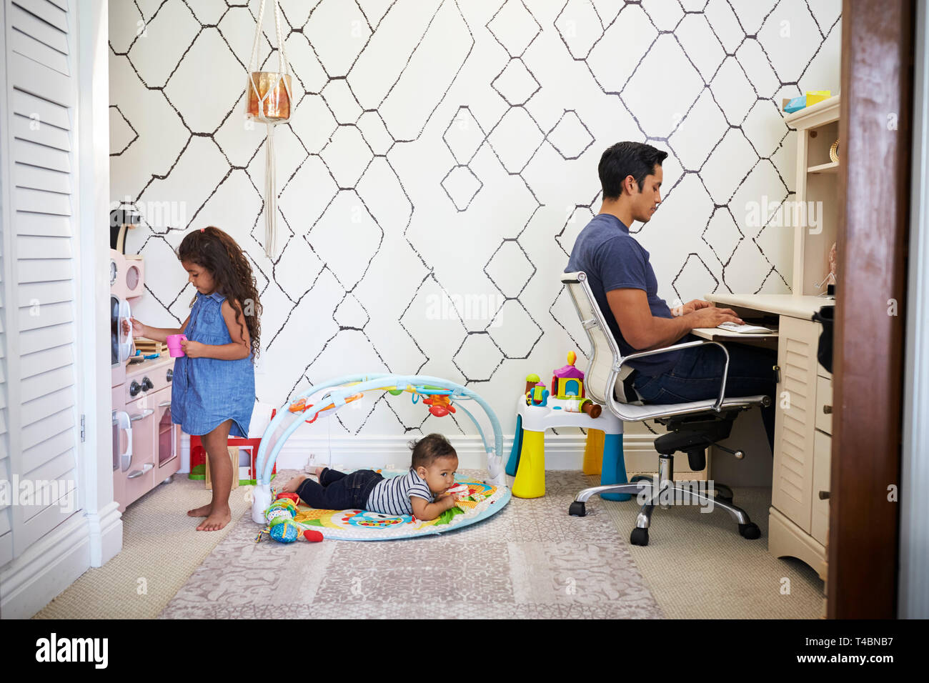 Dad sits working at a desk at home while his baby son and young daughter play in the room behind him Stock Photo