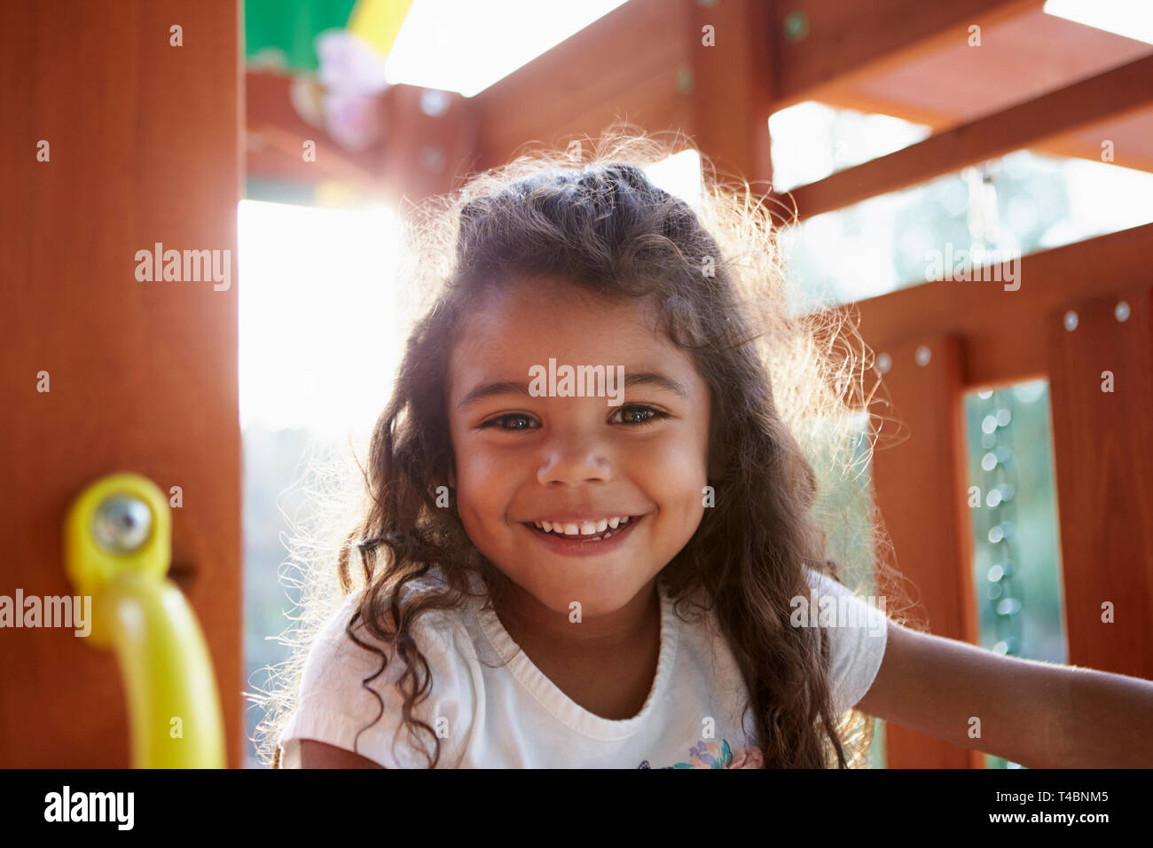 Young Hispanic girl playing on a climbing frame in a playground smiling to camera, backlit, close up Stock Photo