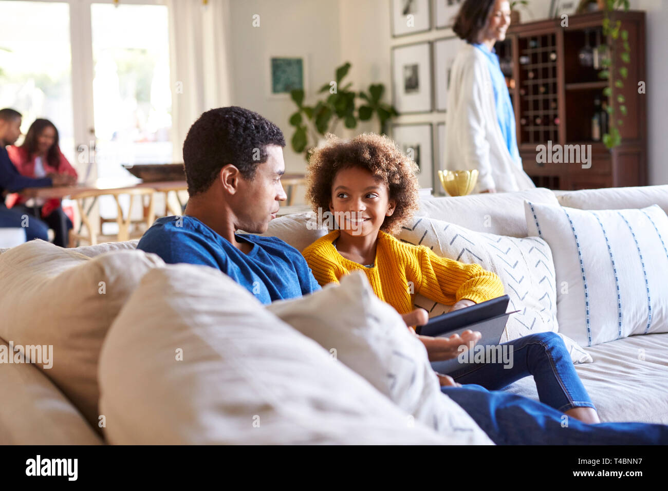 Father spending time with his pre-teen daughter on a sofa in the living room, mother walking through the room, and grandparents sitting at a table in the background Stock Photo