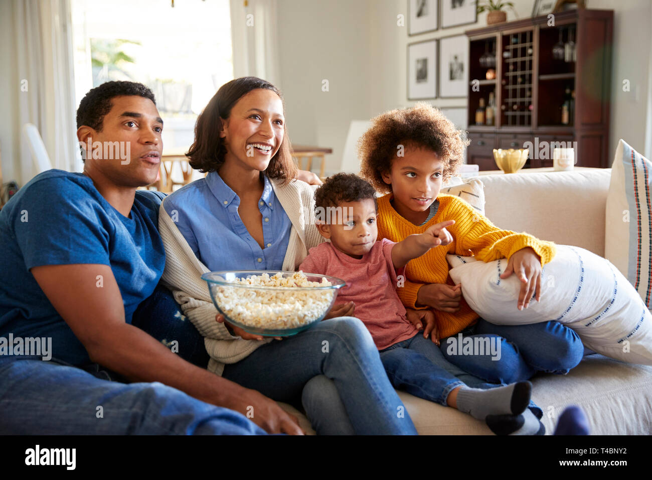 Young family sitting together on the sofa in their living room watching TV and eating popcorn Stock Photo