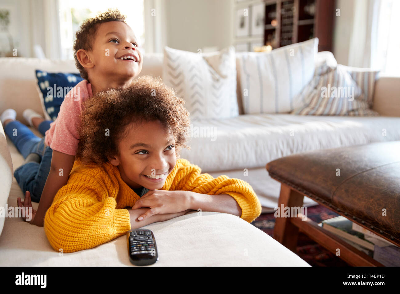 Pre-teen girl lying on sofa watching TV in the living room with her younger brother sitting on her back, close up Stock Photo