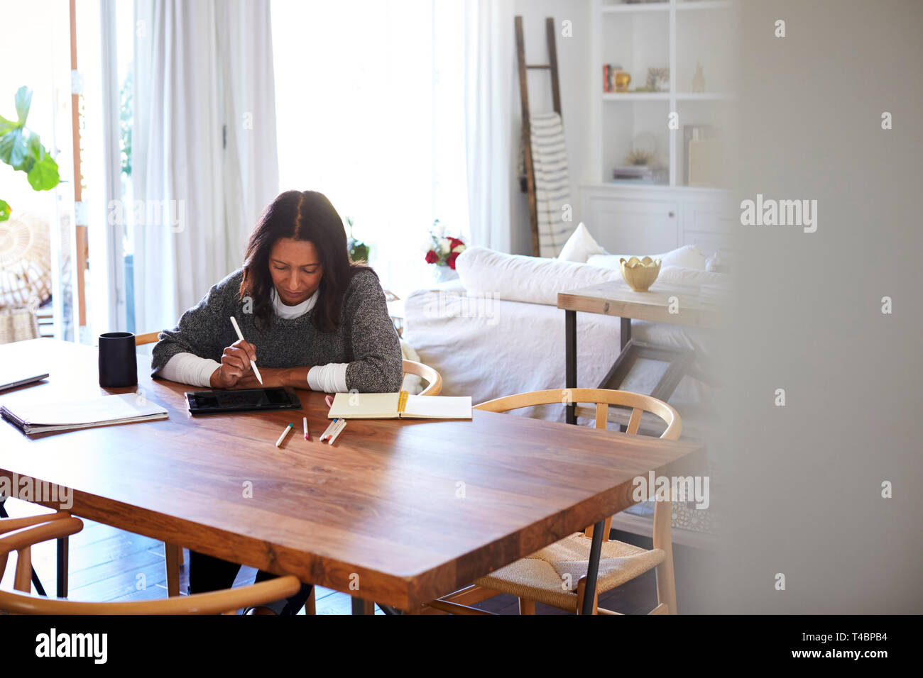 Middle aged mixed race woman sitting at a table in her dining room using a stylus with a tablet computer, seen from doorway Stock Photo