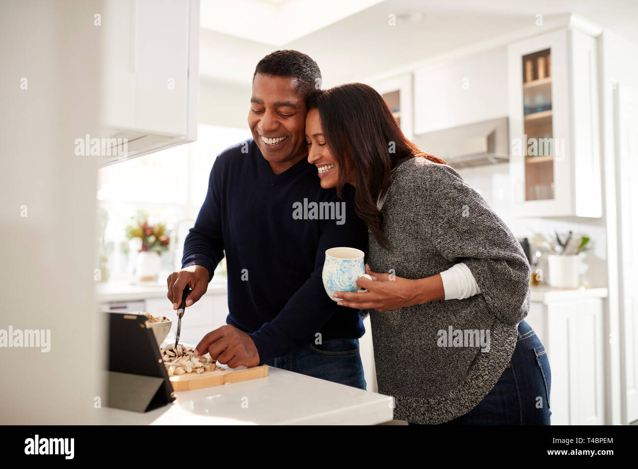 Middle aged mixed race couple standing at worktop in the kitchen preparing food following a recipe on a tablet computer, selective focus Stock Photo