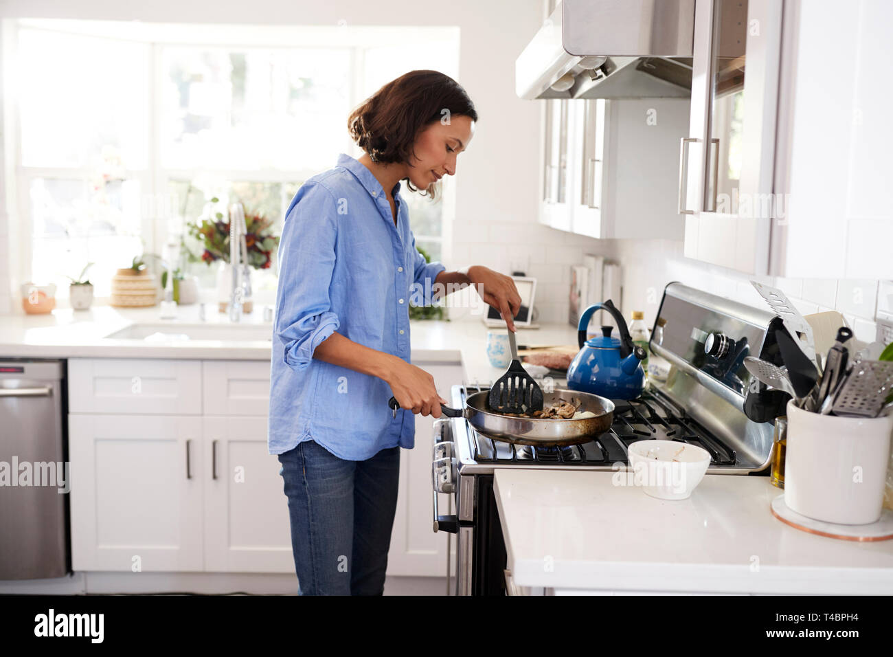 Young adult woman standing at hob in the kitchen cooking food using a spatula and frying pan, side view Stock Photo