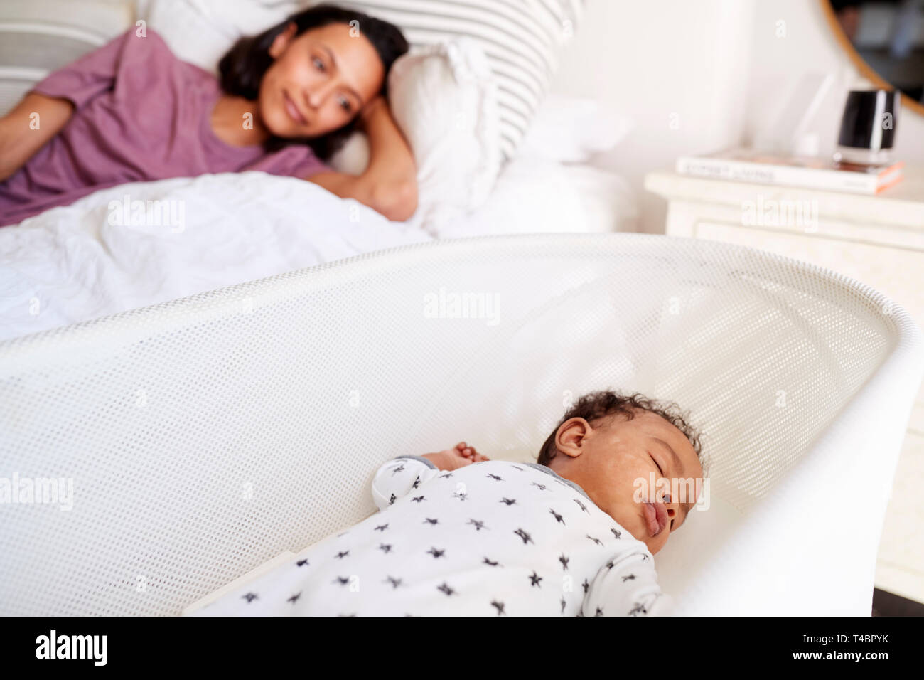 Close up of young adult mother lying on her bed looking down at her three month old baby sleeping in his cot Stock Photo