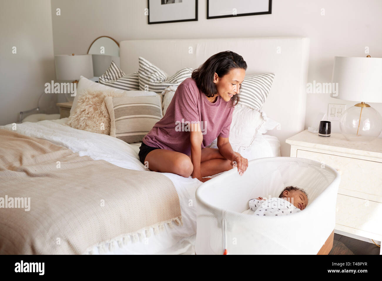 Young adult mother sitting on her bed looking down at her three month old baby sleeping in his cot Stock Photo