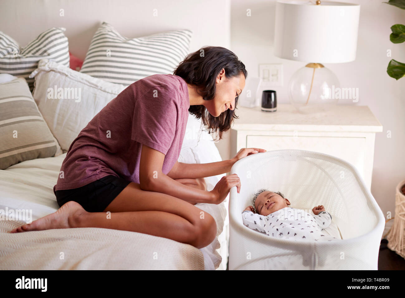 Young adult mother kneeling on her bed looking down at her three month old baby sleeping in his cot, side view Stock Photo