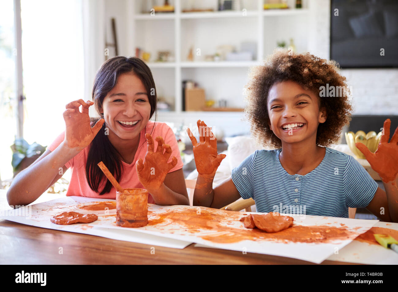 Two girlfriends having fun playing with modelling clay at home, smiling and showing dirty hands to camera, front view, close up Stock Photo