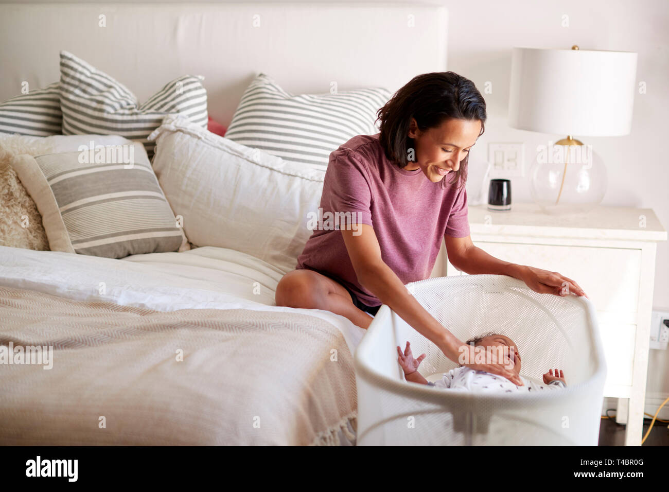 Millennial mixed race mother sitting on her bed comforting her crying baby lying bedside her in his cot Stock Photo