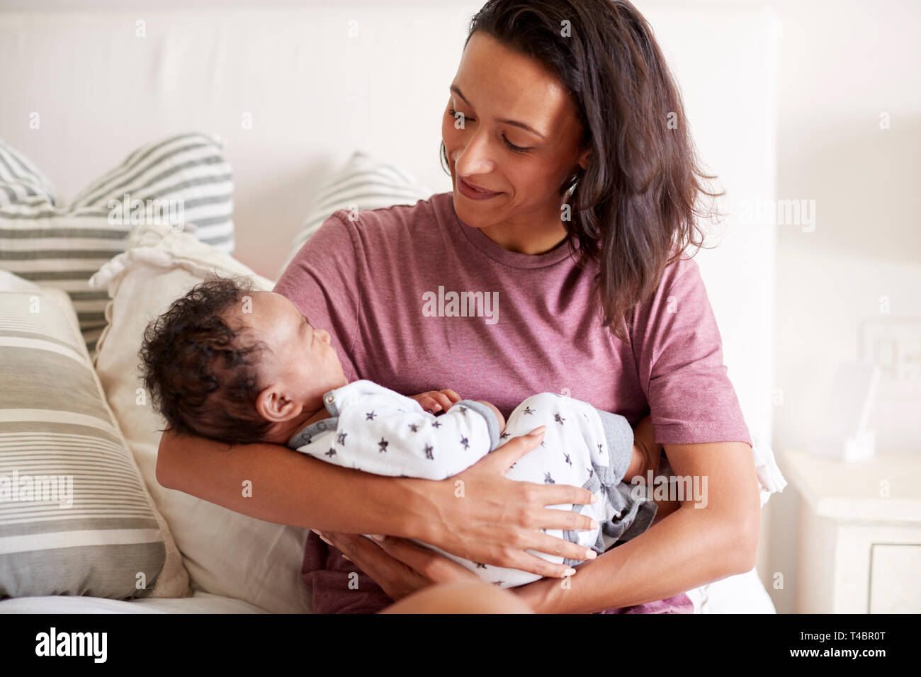 Young mixed race adult mother sitting on her bed and holding her baby son in her arms, waist up, close up Stock Photo
