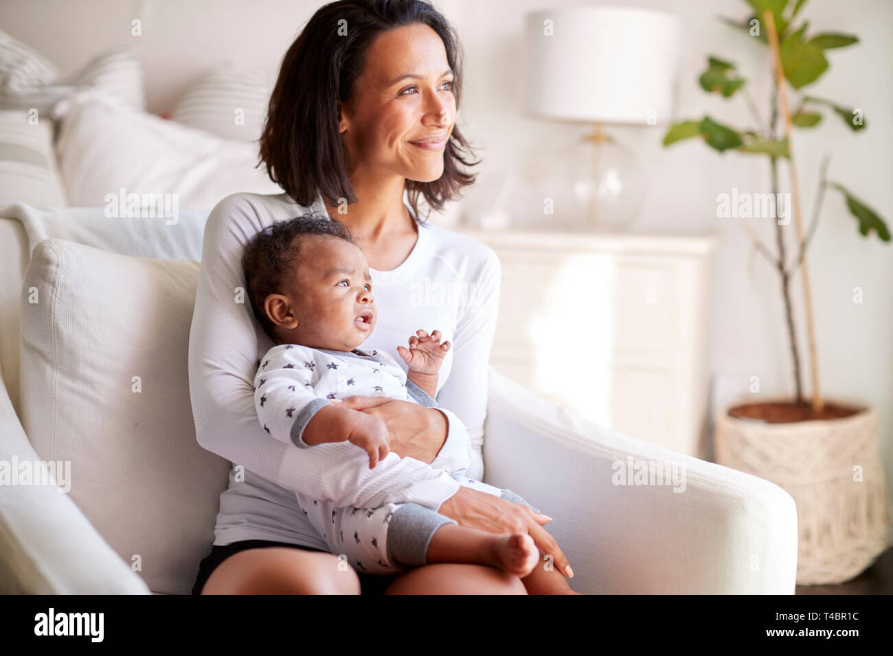 Young adult mother sitting in an armchair in her bedroom, holding her three month old baby son in her arms and looking away smiling, close up Stock Photo