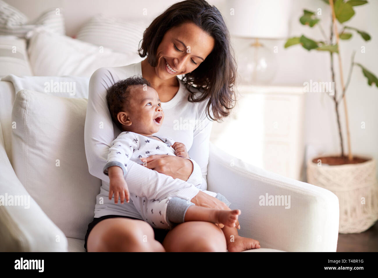 Young adult mother sitting in an armchair in her bedroom, holding her three month old baby son in her arms and looking down at him smiling, close up Stock Photo