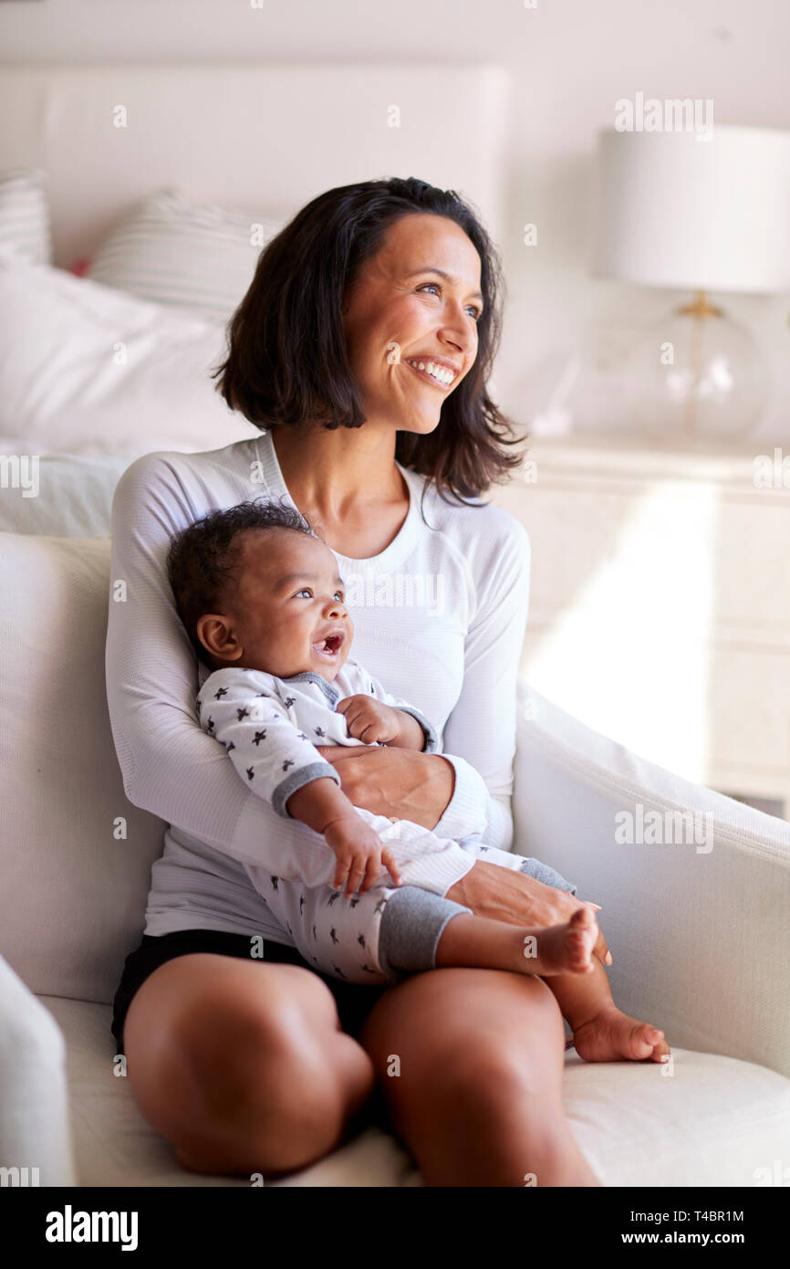 Young adult mother sitting in an armchair in her bedroom, holding her three month old baby son in her arms and looking away smiling, vertical Stock Photo