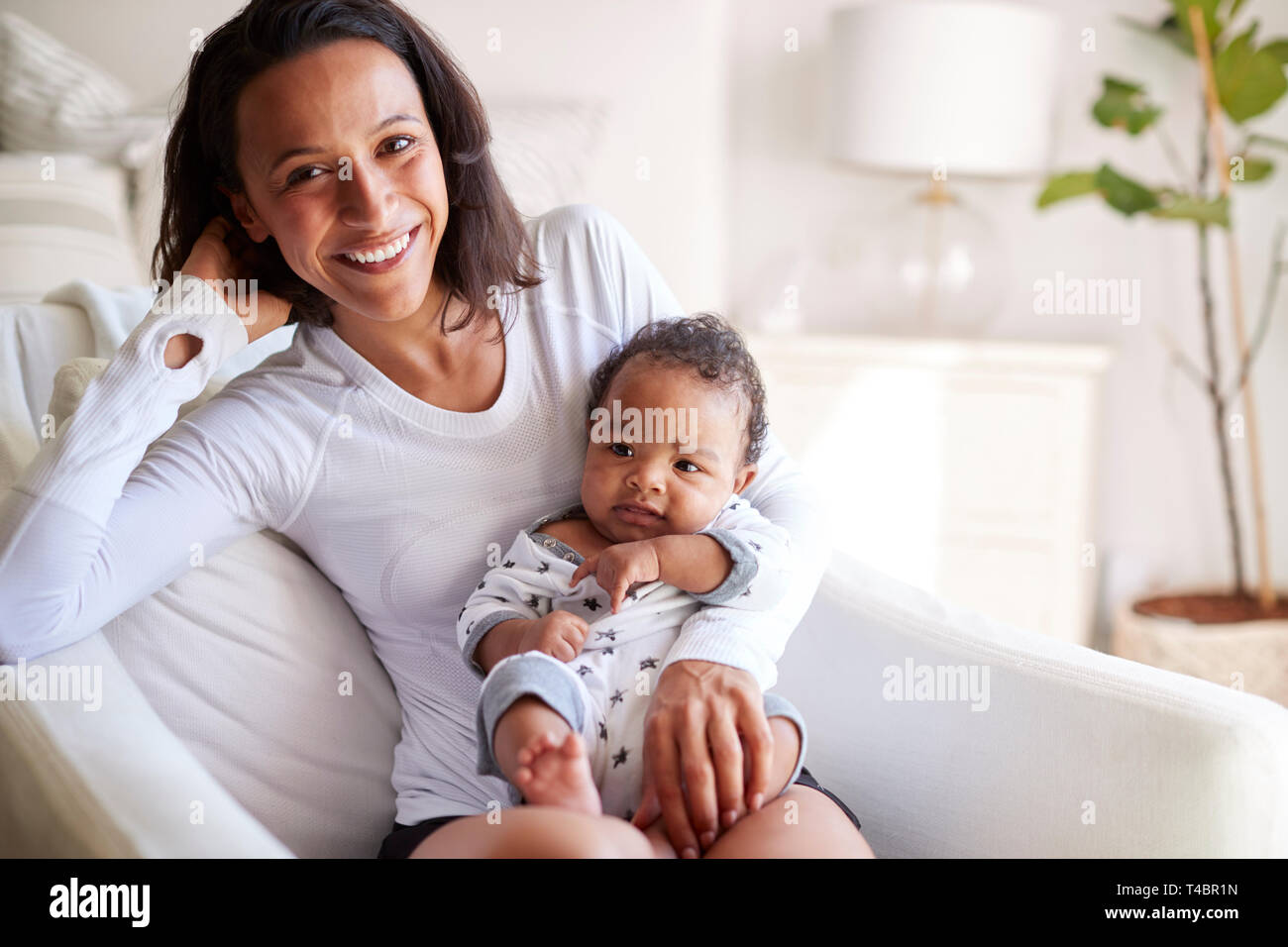 Happy mixed race young adult mother sitting on an armchair holding her three month old baby son, smiling to camera, close up Stock Photo