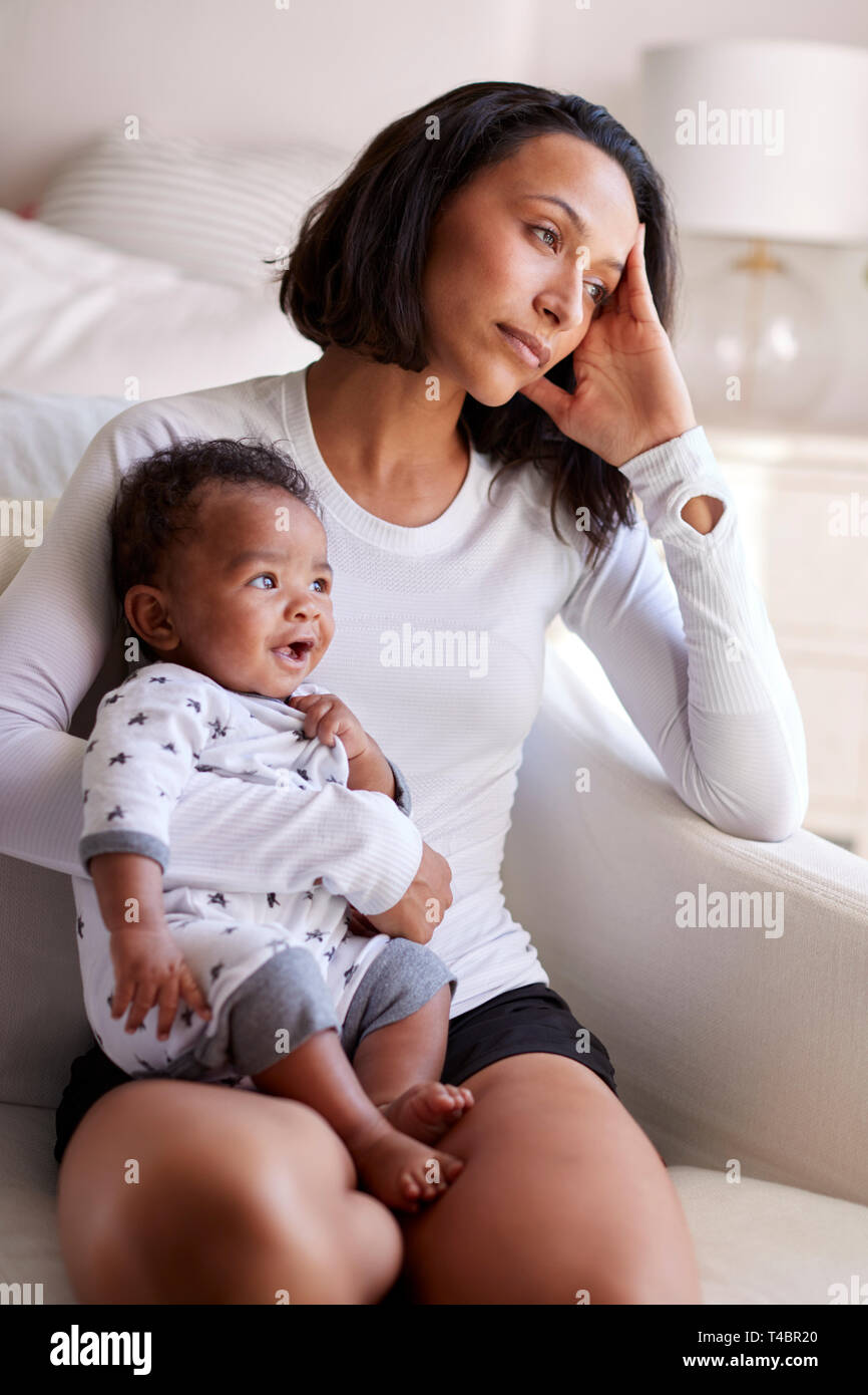 Young adult mother sitting in an armchair in her bedroom, holding her three month old baby son, looking away in contemplation, vertical Stock Photo