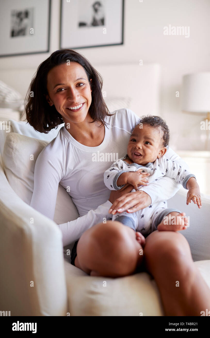 Happy mixed race young adult mother sitting on an armchair holding her three month old baby son, smiling to camera, vertical Stock Photo