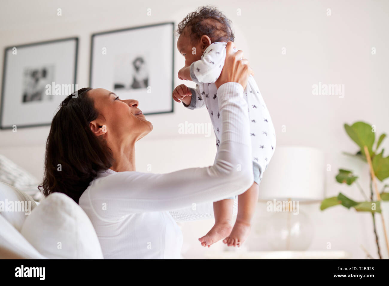 Happy mixed race young adult mother raising her three month old baby son in the air, smiling at him, close up, side view Stock Photo