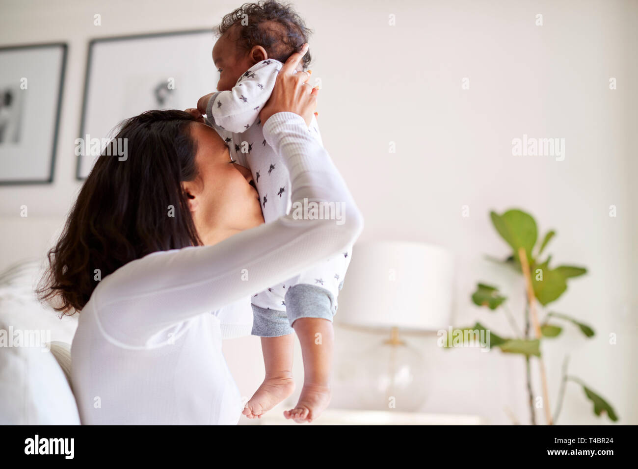 Happy mixed race young adult mother raising her three month old baby son in the air and kissing his tummy, close up, side view Stock Photo